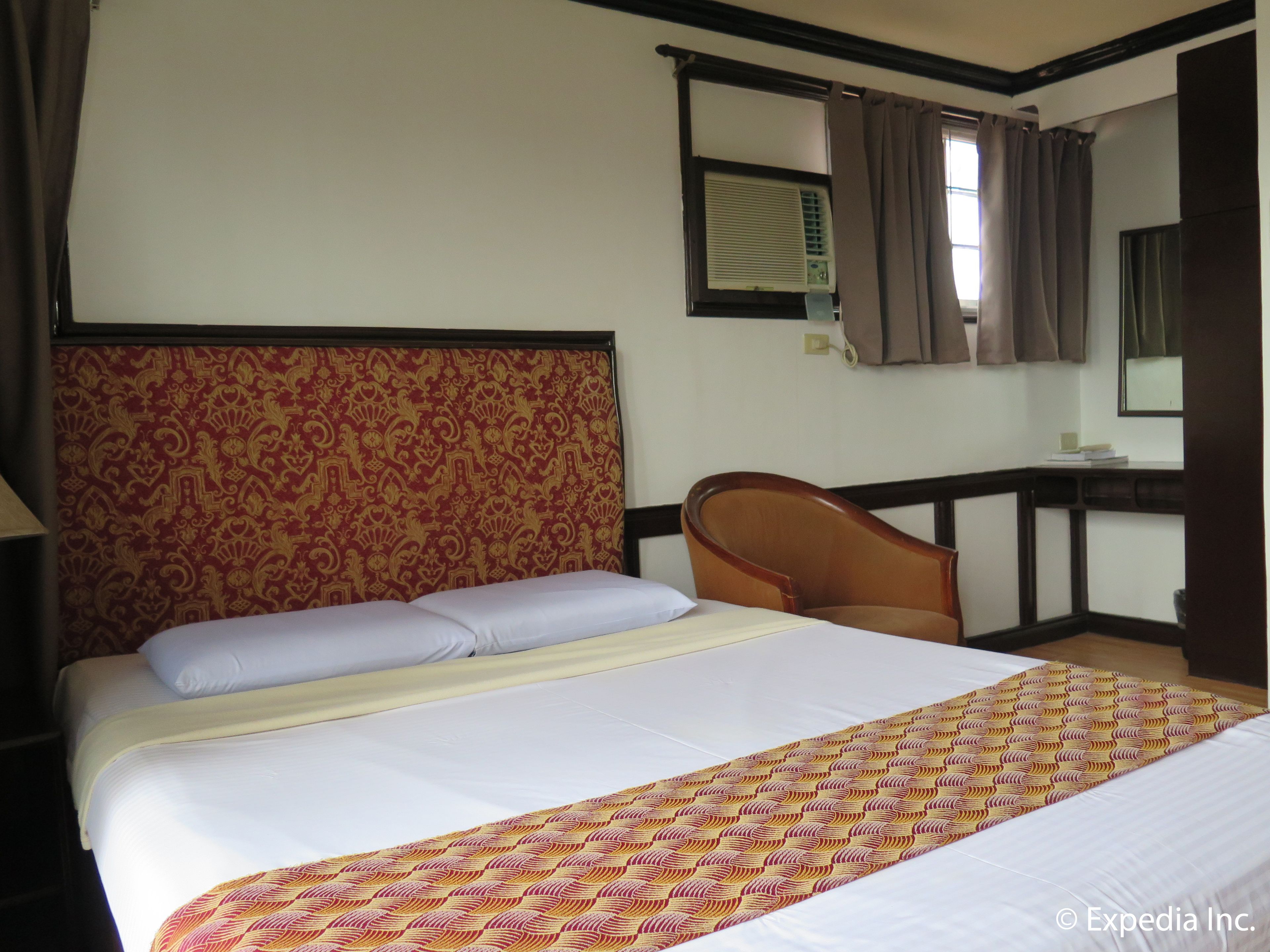 A hotel room with a large bed, a wooden headboard, a desk with a chair, a window with curtains, and a wall-mounted air conditioning unit.