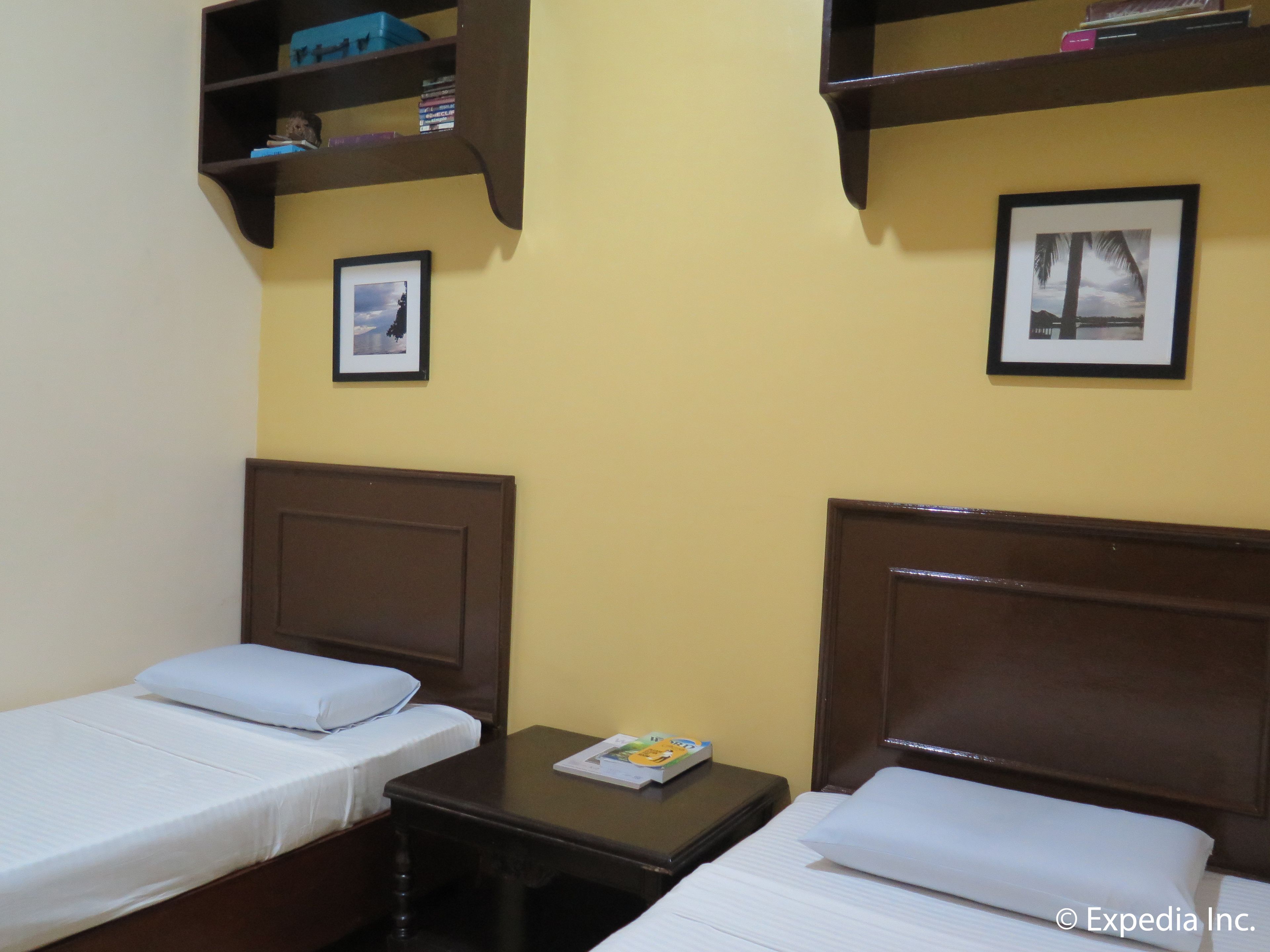 A hotel room with two single beds, a small table with a book, a shelf with books and a picture, and a framed picture on the wall.