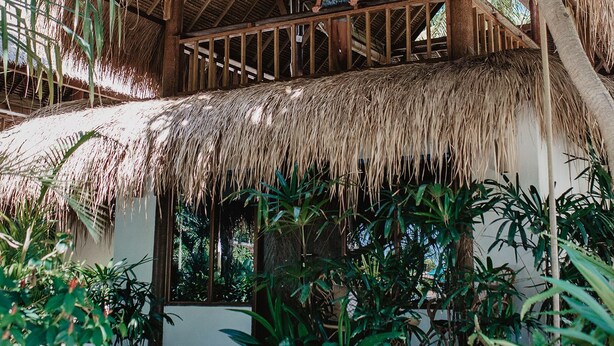 Une construction à toit de chaume, dotée d’un balcon en bois, entourée d’une végétation luxuriante.