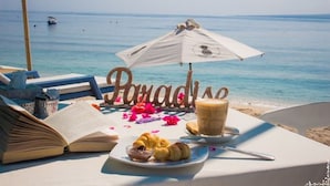 Une table installée sur la plage, avec un livre, des croissants et une boisson, le tout mis en valeur par le décor : la mer en toile de fond et un panneau en bois sur lequel on peut lire le mot « Paradis ».
