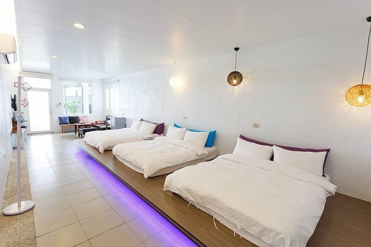 A hotel room with three beds, a desk, and a chair. There is a window providing natural light.