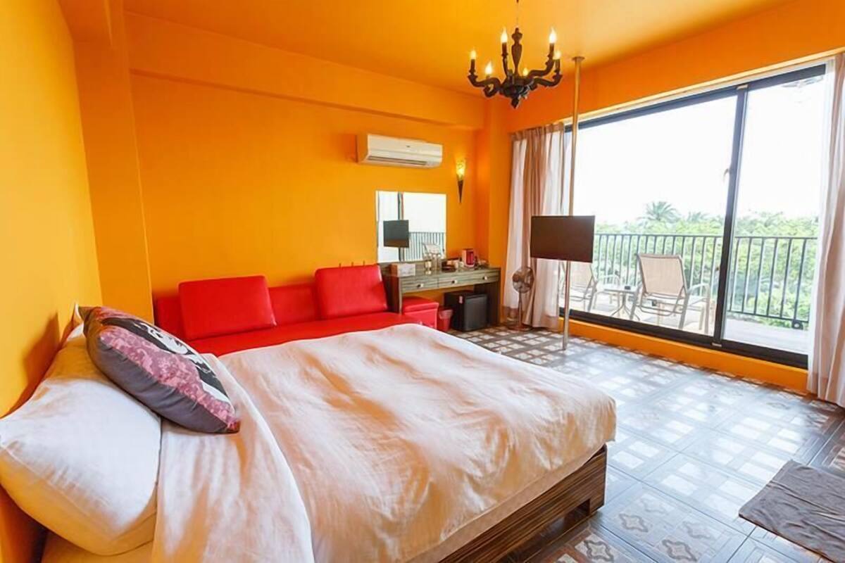 A hotel room with a large bed, a red sofa, a desk with a chair, and a balcony with outdoor furniture.