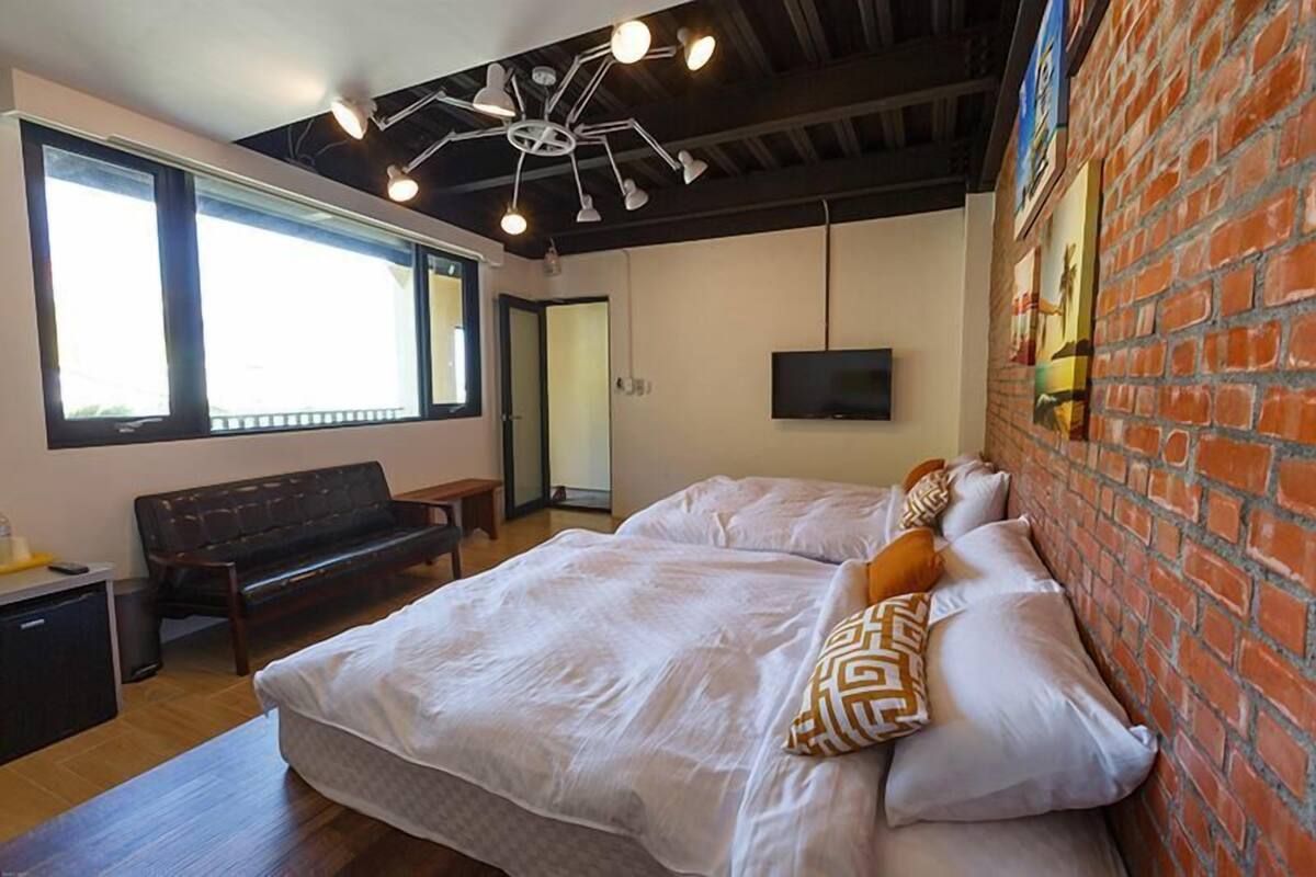 A modern hotel room with a large bed, a bench, a desk, and a TV mounted on the brick wall.