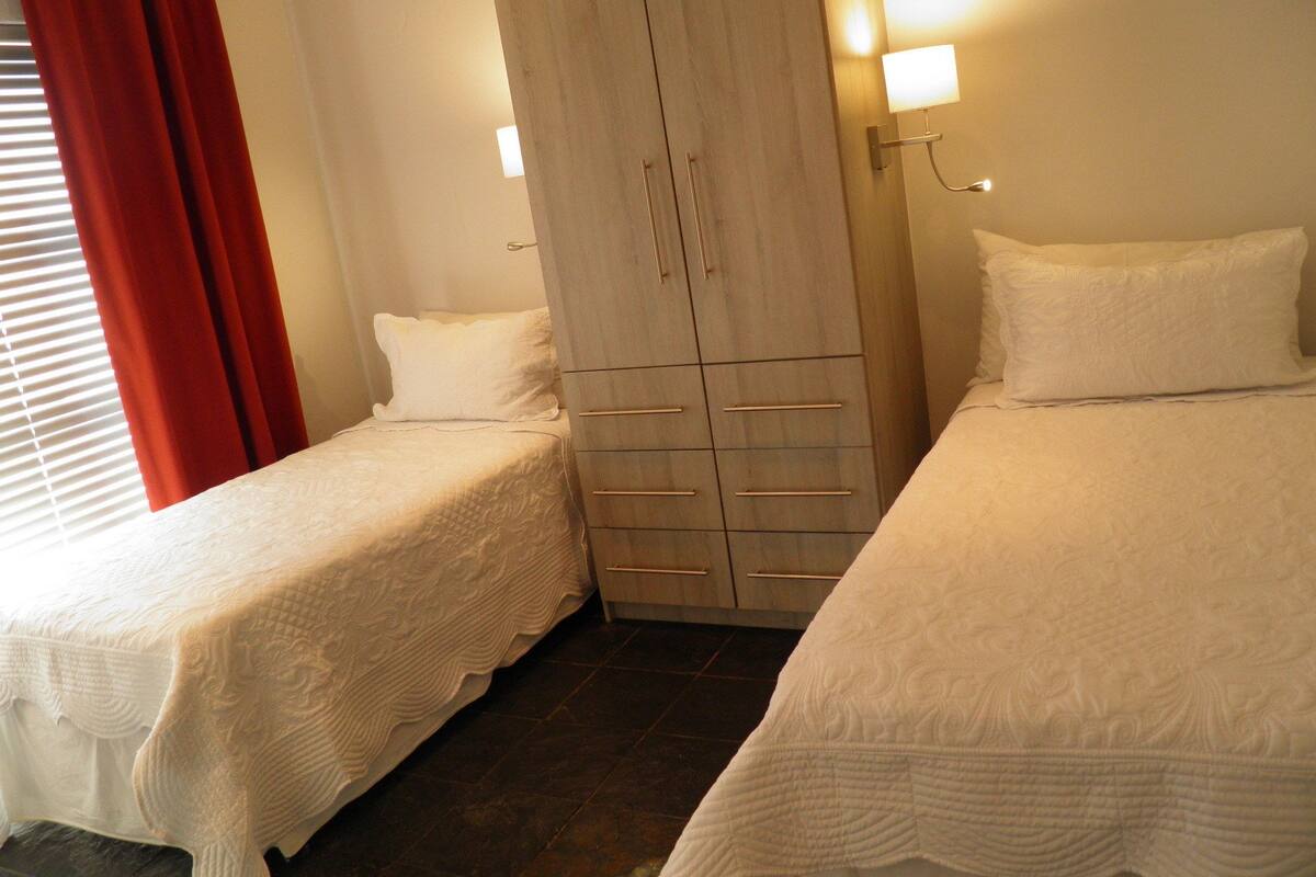 A room with two single beds, a wardrobe, and a window with red curtains.
