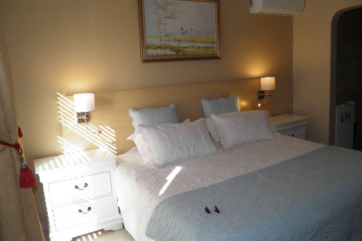 A neatly made bed with white linens and pillows, a painting above the headboard, and a wall-mounted air conditioner.