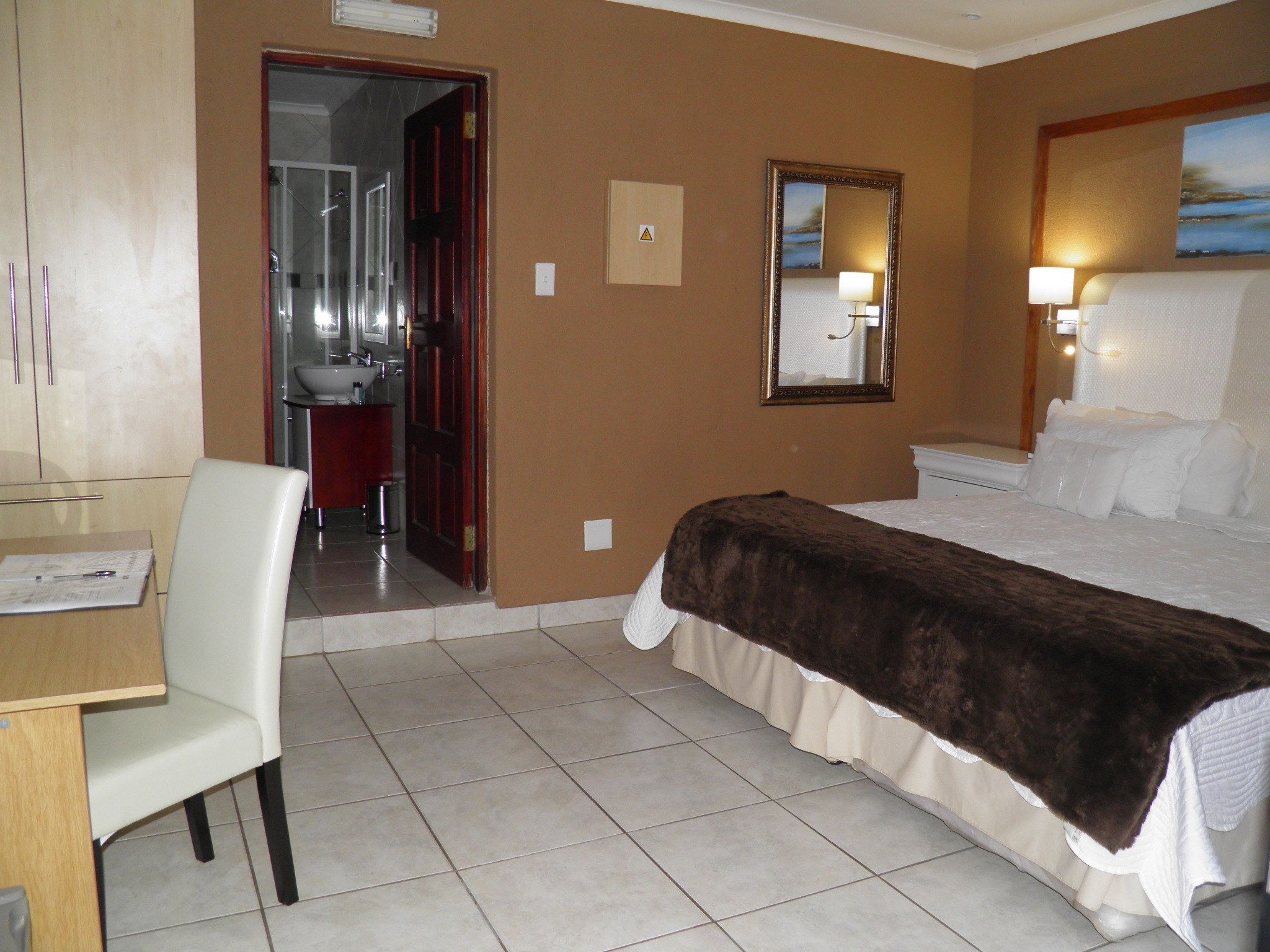 A hotel room with a bed, a desk with papers, a chair, a bathroom visible through an open door, and a painting on the wall.