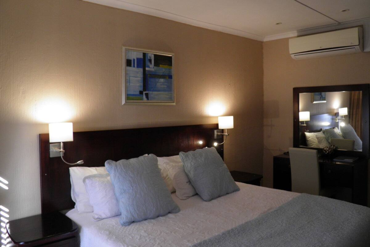 A hotel room with a bed, two bedside lamps, a nightstand, a mirror, and an air conditioning unit.