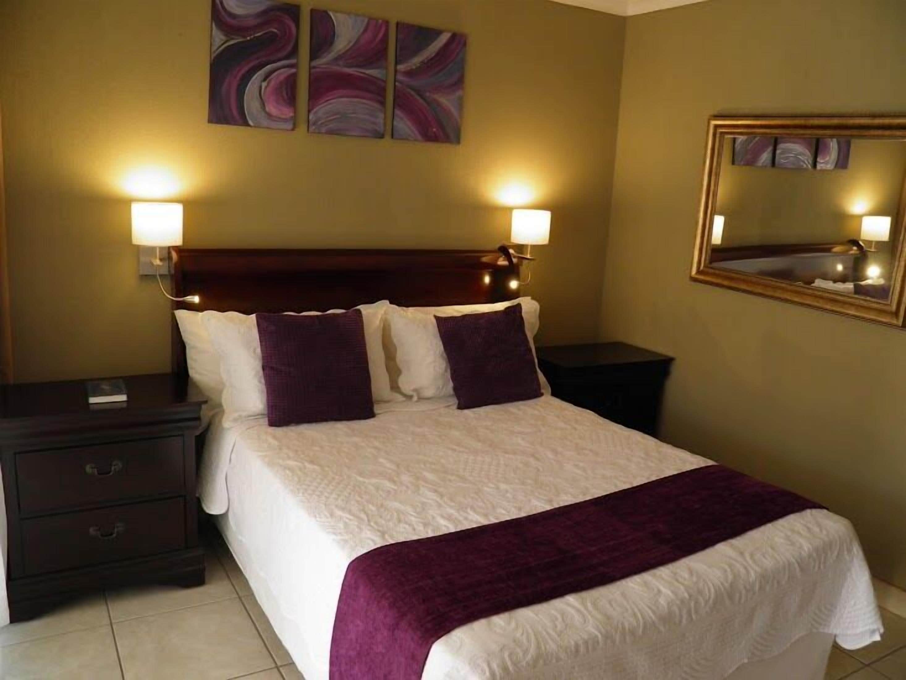 A neatly made bed with a white and purple bedspread, two bedside lamps, a dark wooden nightstand, and a wall-mounted mirror.
