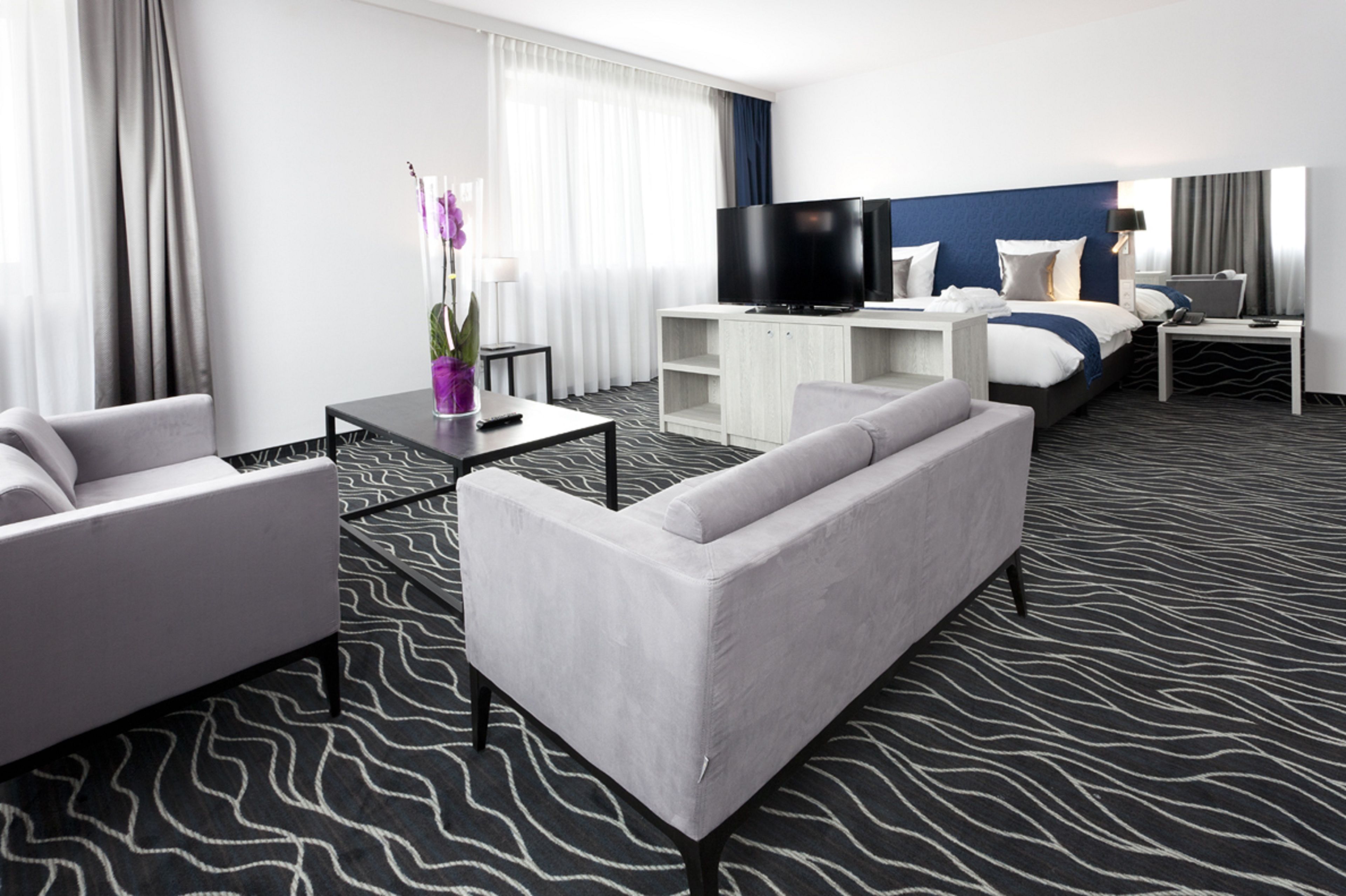 A modern hotel room with a sofa, a TV, a bed with a blue headboard, and a patterned carpet.