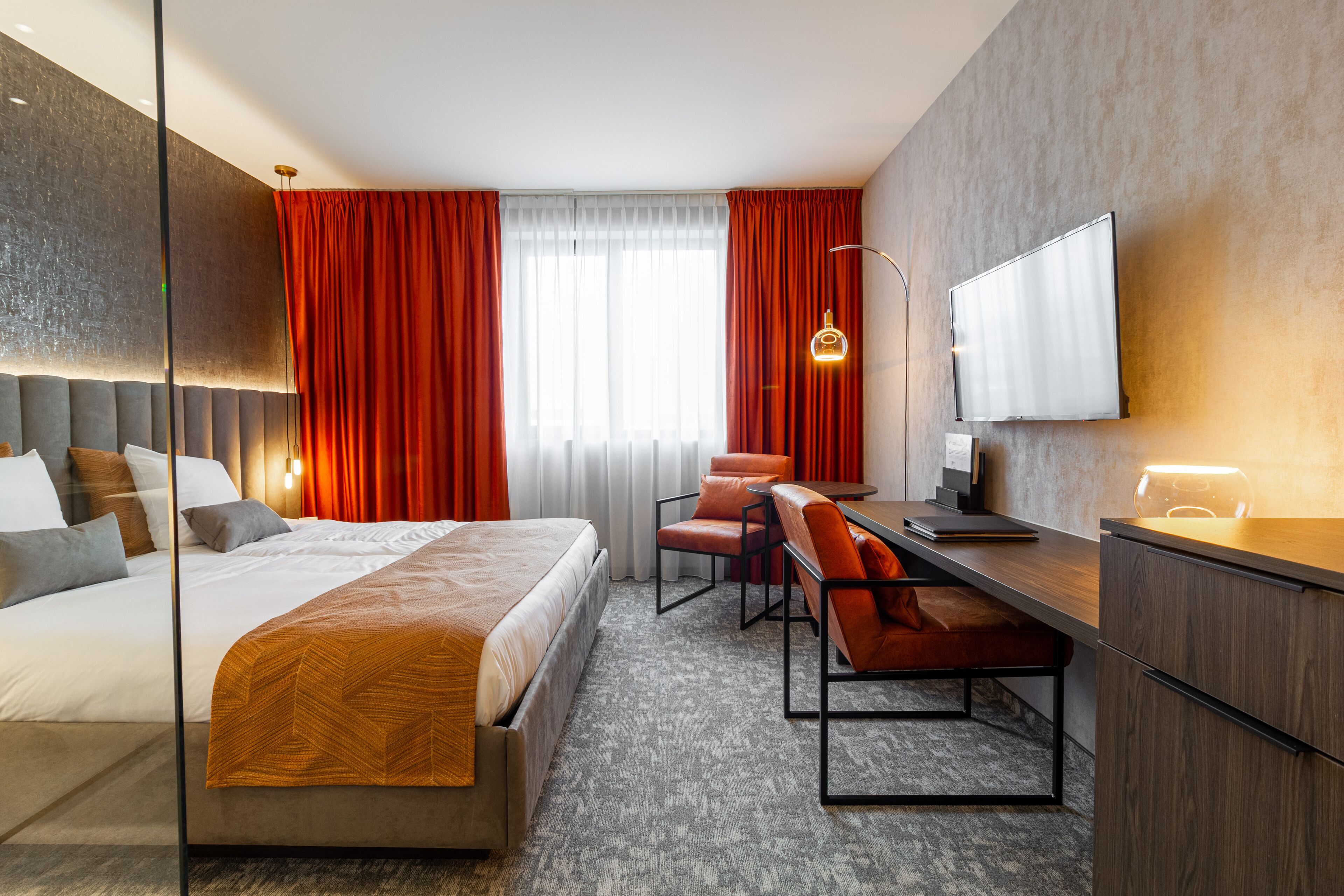 Ein modernes Hotelzimmer mit einem großen Bett, einem Schreibtisch mit Stuhl, einem Flachbildfernseher und einem großen Fenster mit roten Vorhängen.