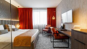 Ein modernes Hotelzimmer mit einem großen Bett, einem Schreibtisch mit Stuhl, einem Flachbildfernseher und einem großen Fenster mit roten Vorhängen.