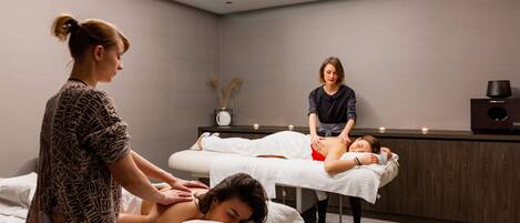 Un cuarto de masoterapia con dos clientes recibiendo masajes, un terapeuta atendiéndolos y un área de recepción con un reloj y plantas decorativas.