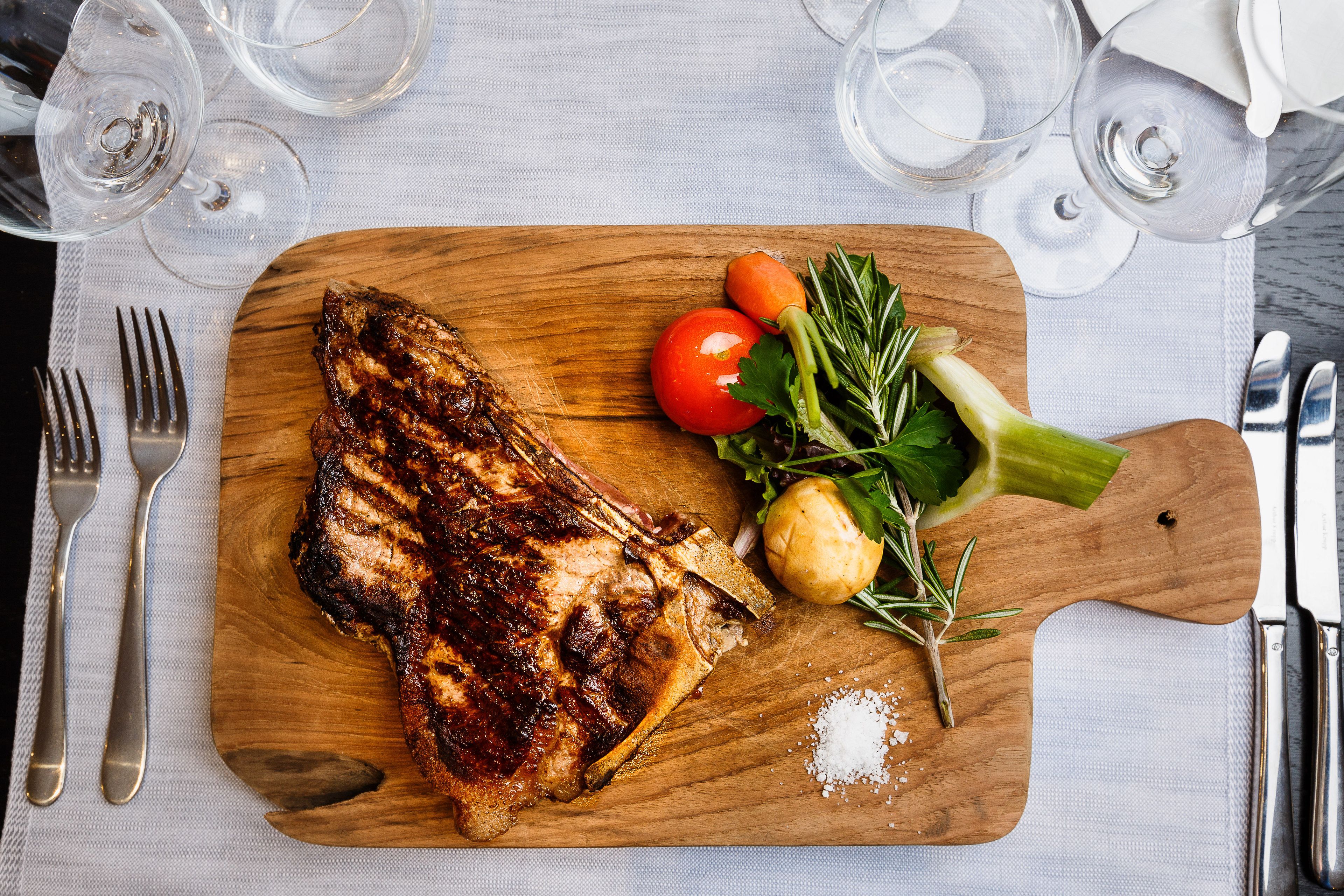 Une planche à découper en bois sur laquelle on voit un steak grillé, une tomate, une carotte et quelques herbes.