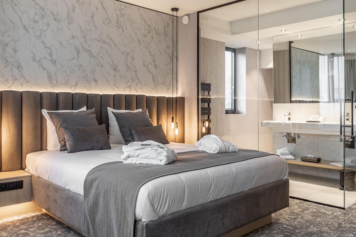 A modern bedroom with a large bed, a bedside table, a built-in bench, a mirror, and a bathroom with a sink and mirror.