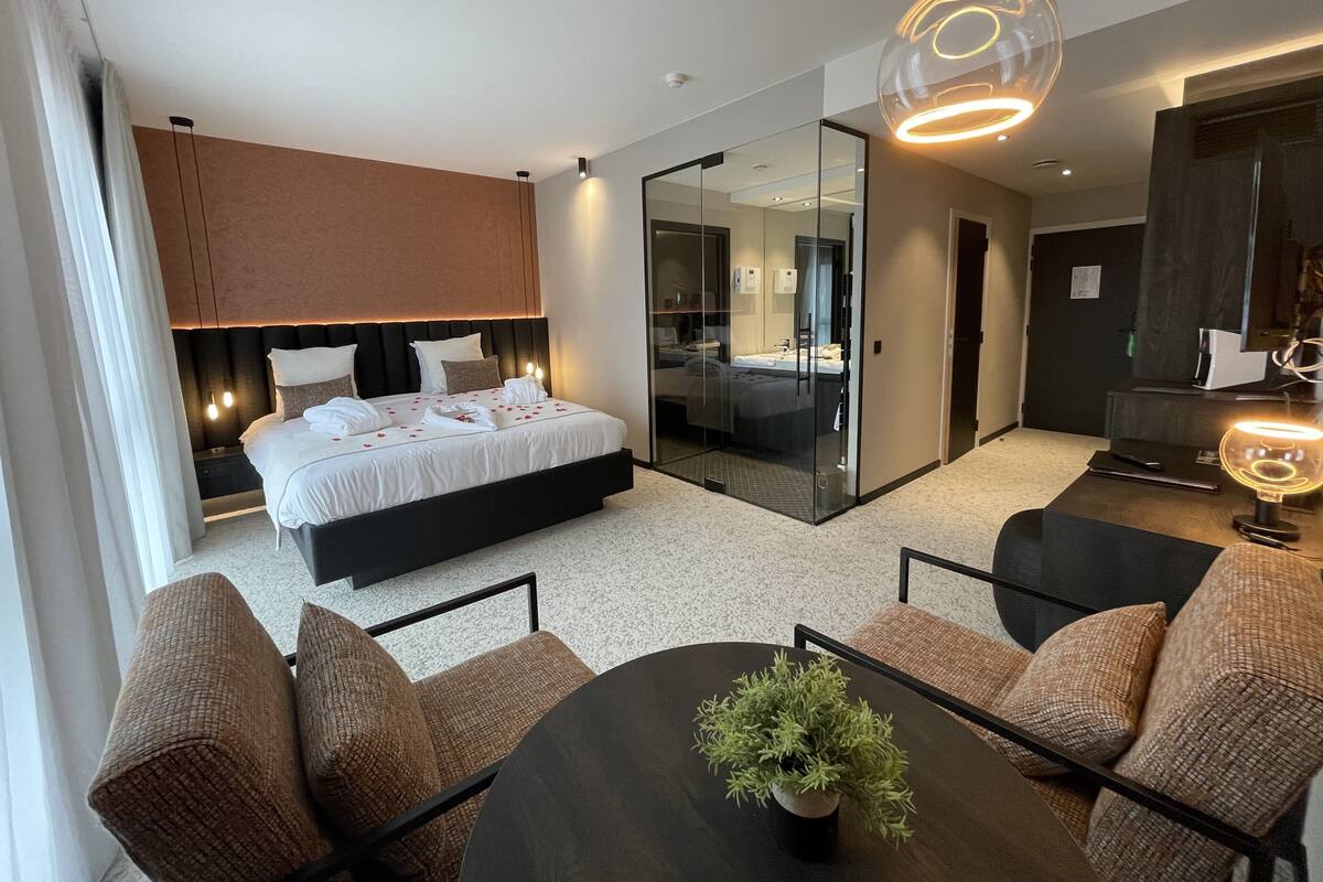 A modern hotel room with a large bed, a glass partition, a dining area, and a small plant on the table.