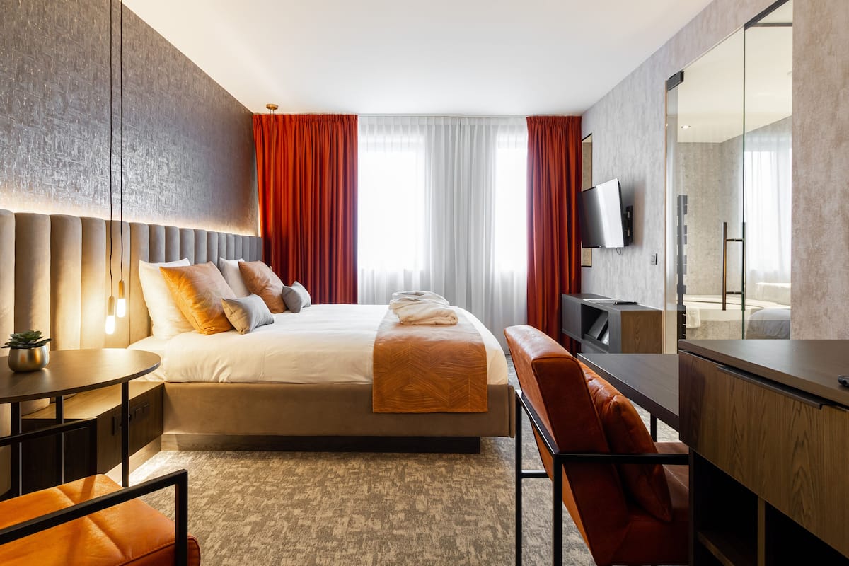 A modern hotel room with a large bed, a desk, a chair, a TV, and a large mirror.
