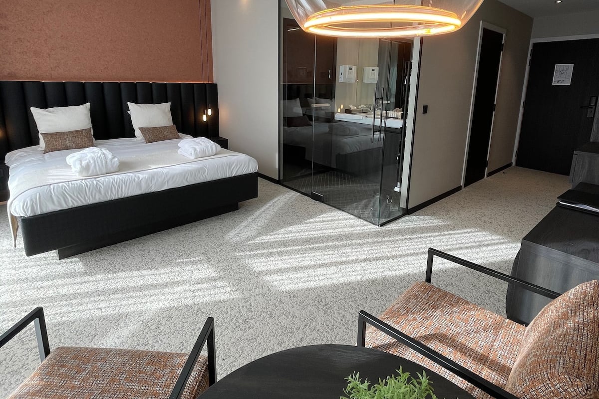 A modern hotel room with a large bed, a glass-enclosed bathroom, a dining area with a table and chairs, and a unique pendant light fixture.