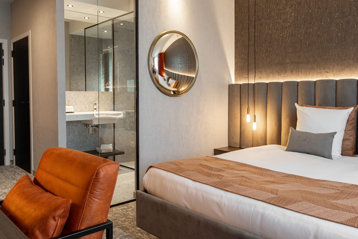 A modern hotel room with a large bed, a glass shower enclosure, and a seating area with a leather chair.