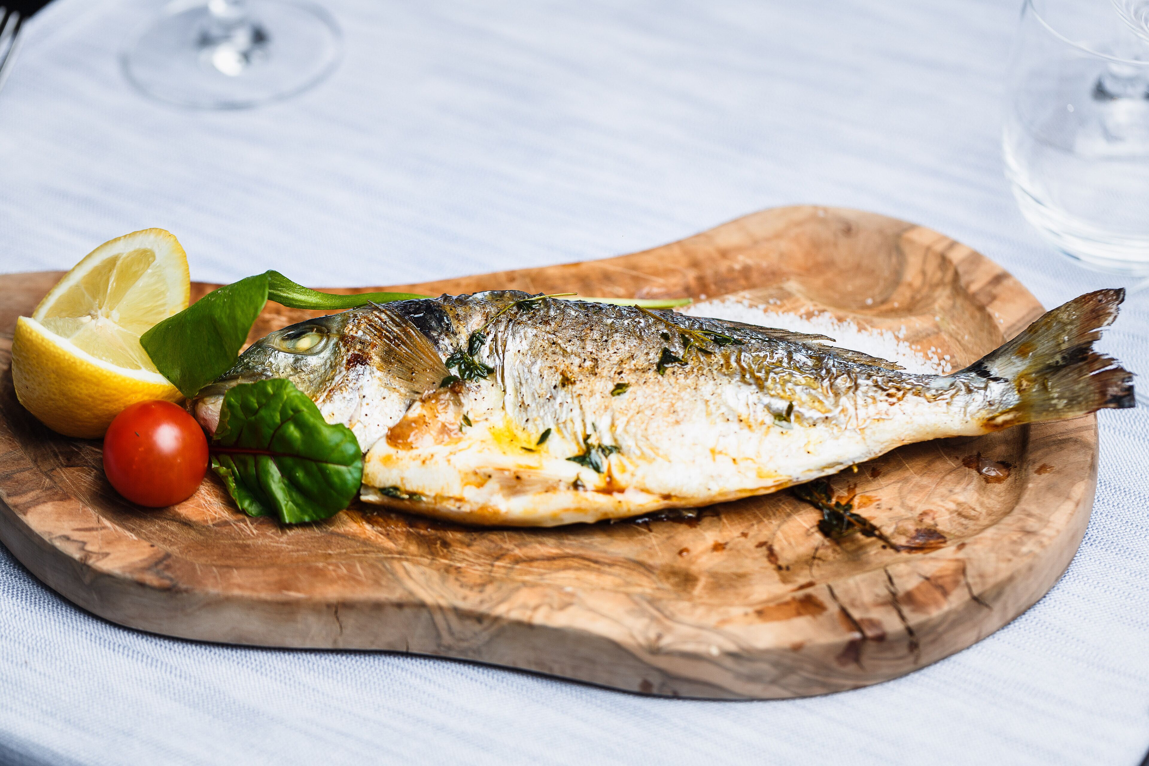 Un poisson grillé, servi sur une planche en bois, accompagné de citron, de tomates et de basilic.