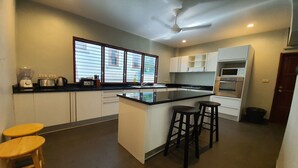 A modern kitchen with a central island, white cabinets, and a built-in oven. There are two stools and a window providing natural light. - NB Villa Celina (Koh Samui)