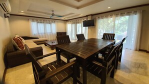A spacious living room with a large wooden dining table, comfortable seating, a ceiling fan, and large windows with curtains. - NB Villa Celina (Koh Samui)