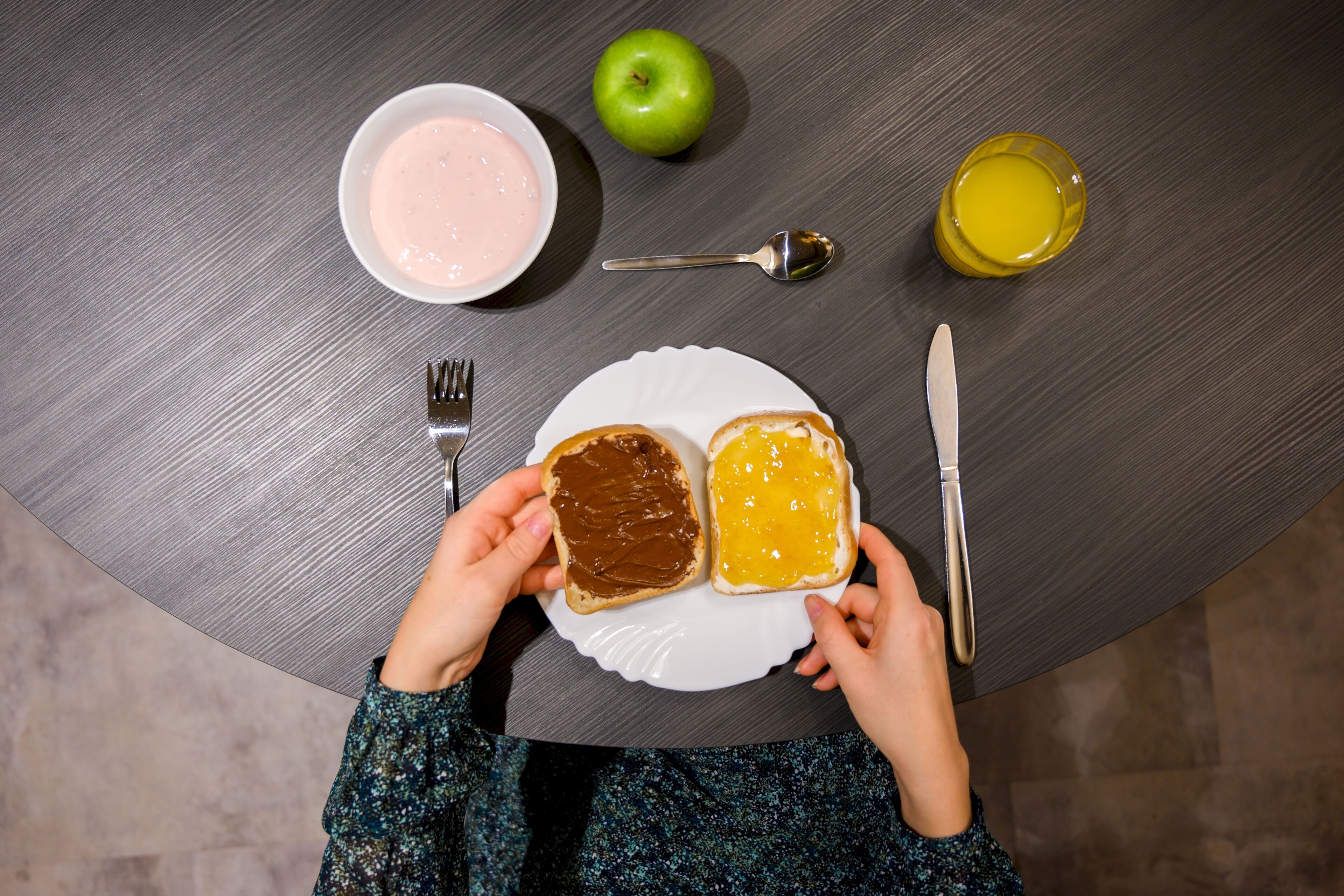A table with a plate of toast, a bowl of yogurt, an apple, a glass of juice, a spoon, a fork, and a knife.