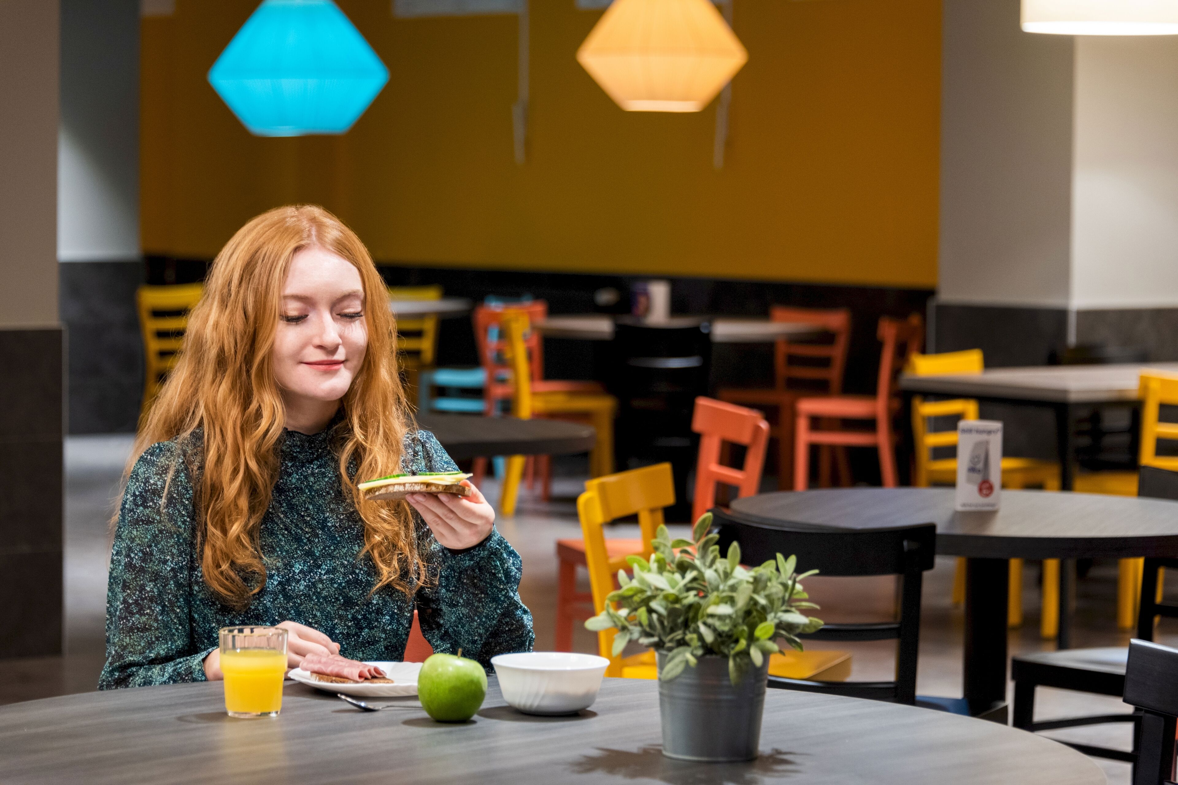 Una persona sentada a una mesa con comida, un vaso de zumo de naranja, una manzana verde y una planta en maceta.