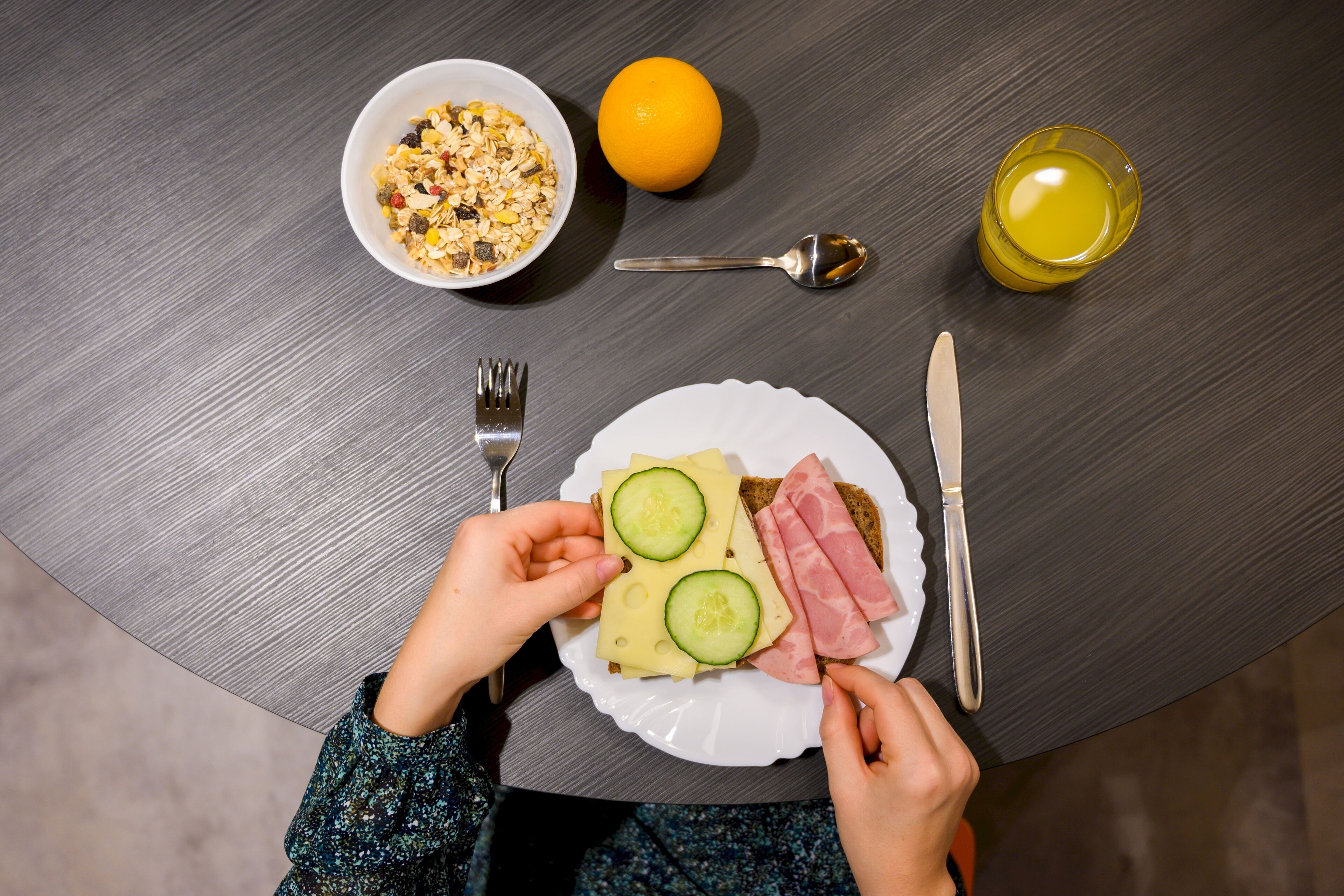 A breakfast setting with a plate of toast, cucumber slices, and ham, a bowl of cereal, an orange, and a glass of juice on a wooden table.