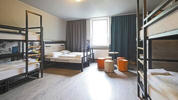 A dormitory room with bunk beds, a window with curtains, a radiator, and a small table.