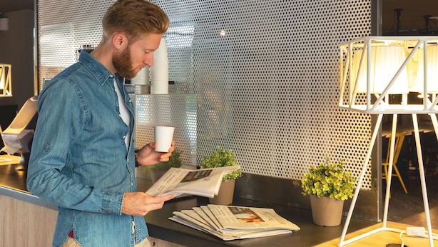 A man reading a newspaper at a counter with a perforated metal wall and a potted plant in the background. - a&o Stuttgart City - Hostel (Stuttgart)