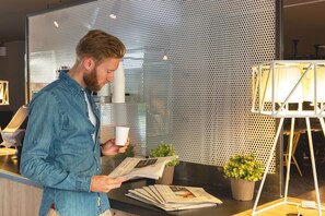 Un homme est assis à un comptoir, lisant un journal. En arrière-plan, on aperçoit un mur en métal perforé et un pot de fleurs.