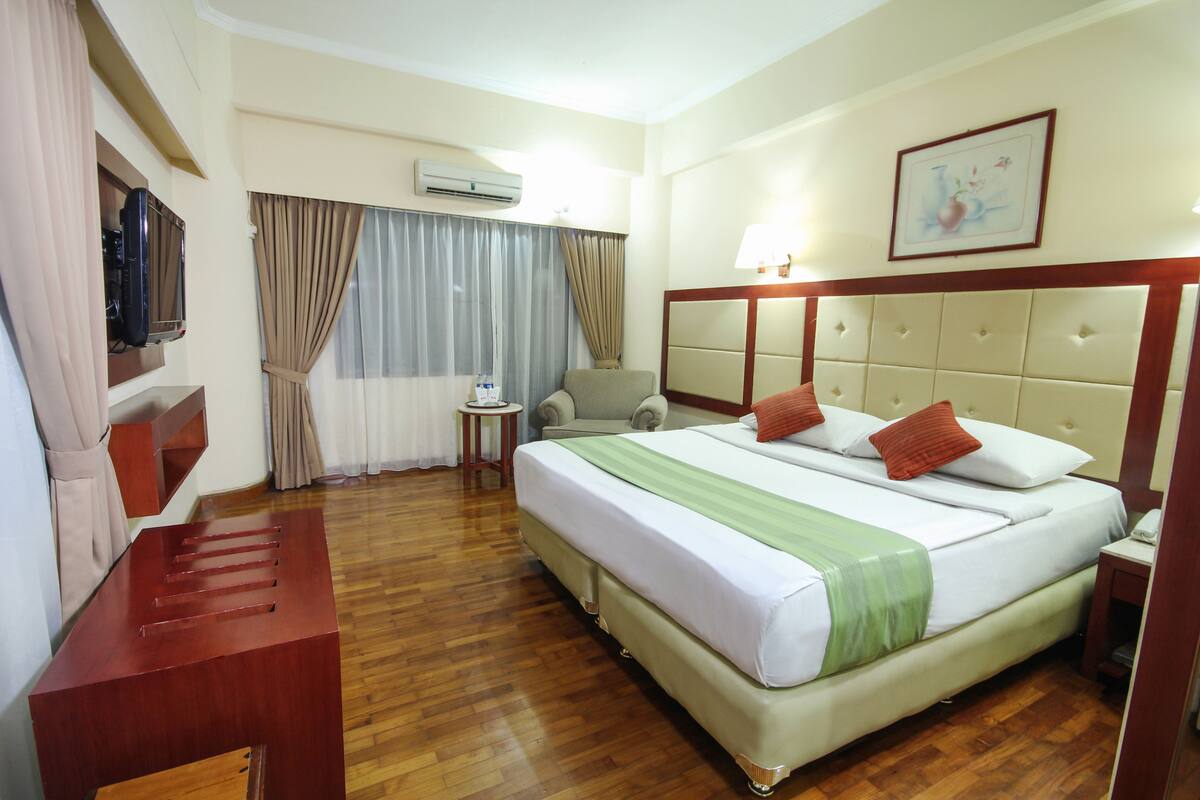 A hotel room with a large bed, a desk, a chair, a TV, and a window with curtains.