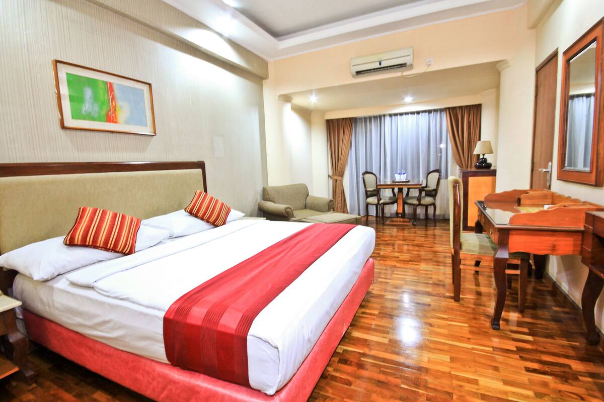 A hotel room with a large bed, a desk, a chair, and a dining table.