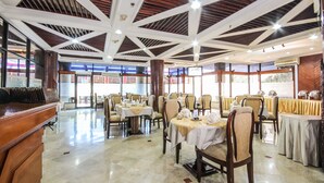 Un'ampia sala da pranzo con soffitti alti, travi in legno e pavimento in marmo lucido. I tavoli sono apparecchiati con tovaglie bianche e sedie.