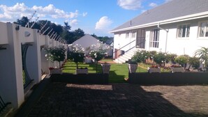 A courtyard with a paved walkway, potted plants, and a white building with a grey roof. - Magaliqua Guest House (Krugersdorp)