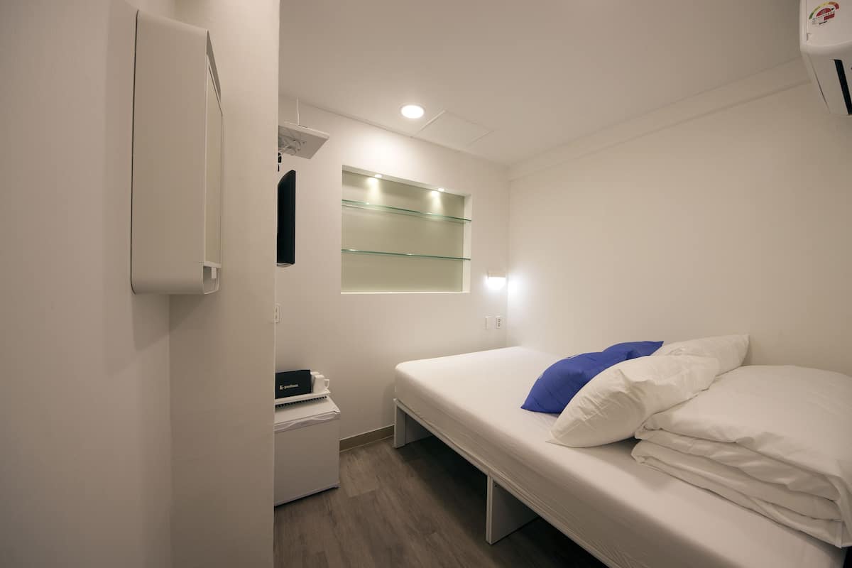 Une petite chambre à coucher, bien rangée, avec un lit simple, une table de chevet, un miroir et une petite étagère.