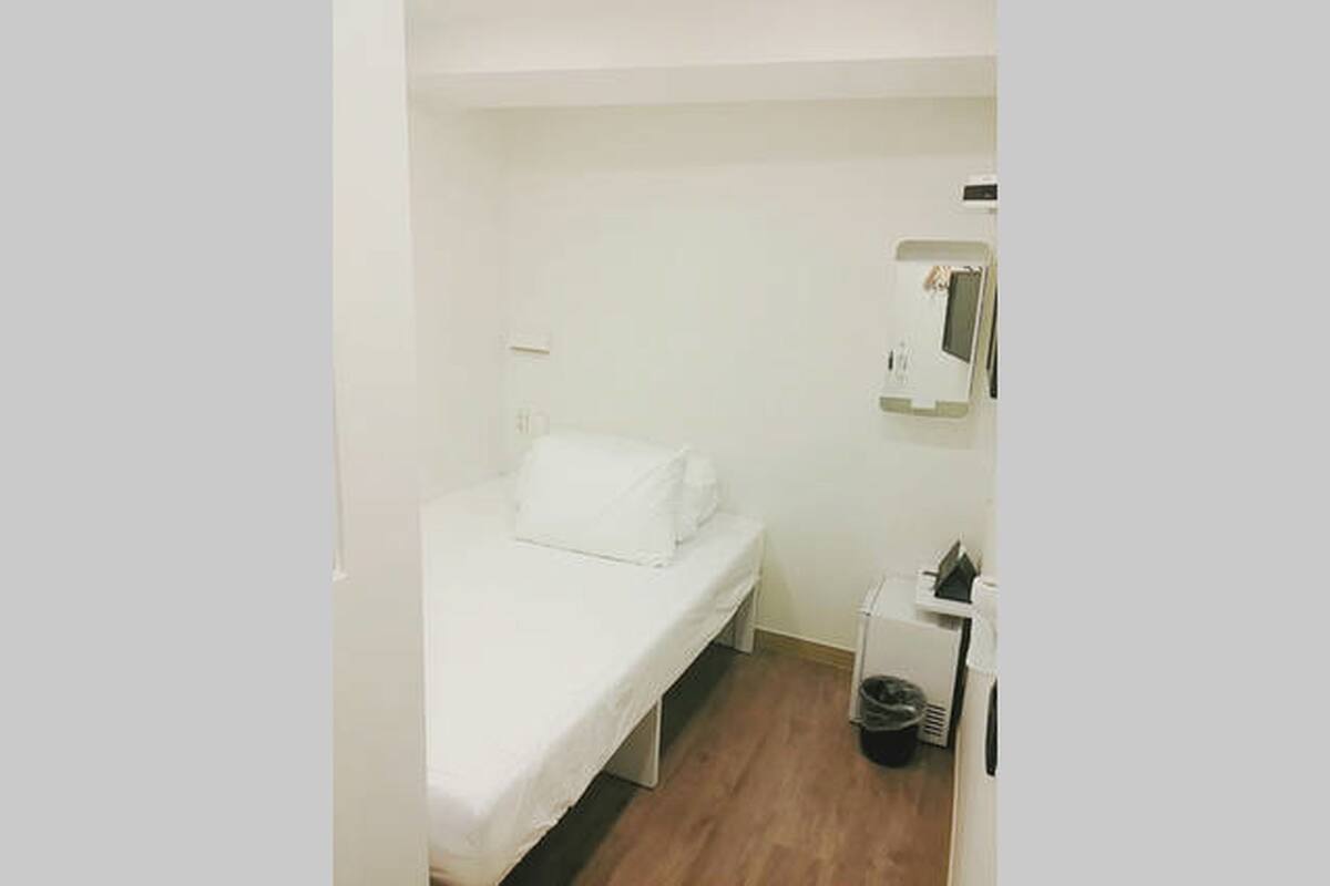 Une petite chambre avec un lit simple, un lit blanc, une petite table de chevet, un miroir fixé au mur et un radiateur.