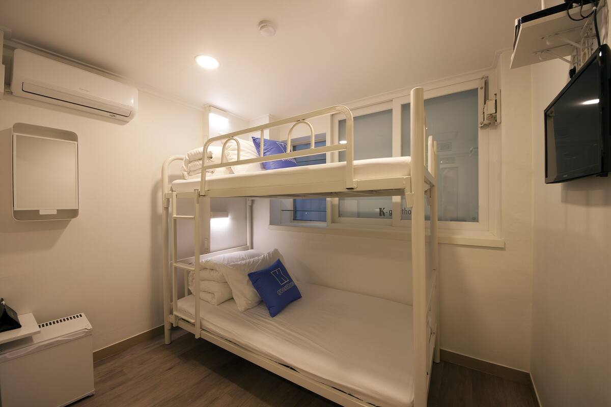 Une chambre avec un lit superposé, décorée dans des tons de blanc, dotée d’une petite fenêtre et d’un téléviseur fixé au mur.