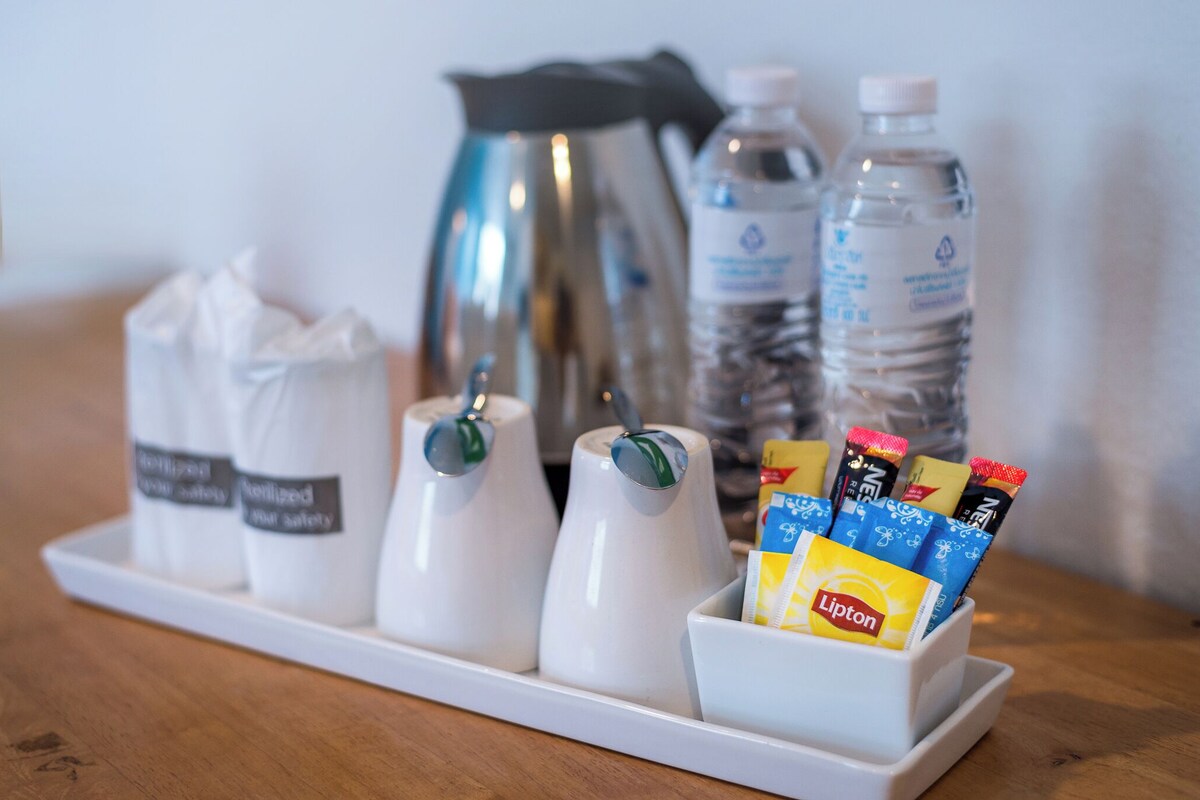 Uma bandeja com diversos artigos de higiene, incluindo uma caixa de lenços, uma cafeteira, garrafas de água e um recipiente com sachês de chá Lipton.
