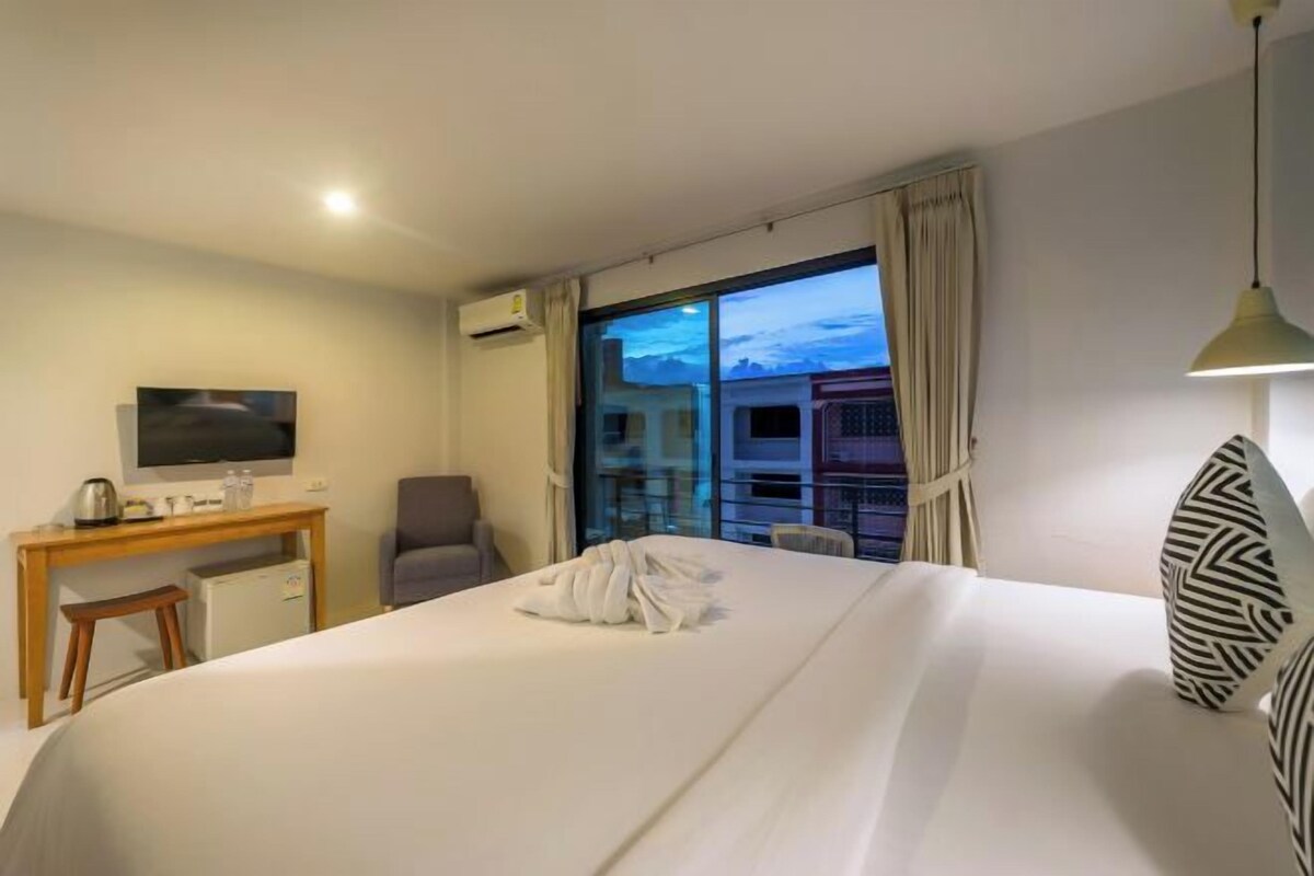 Habitación de hotel con cama, escritorio con televisor, una silla y vista a edificios a través de un ventanal grande.
