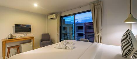 A hotel room with a bed, a desk with a television, a chair, a small refrigerator, and a window with curtains.