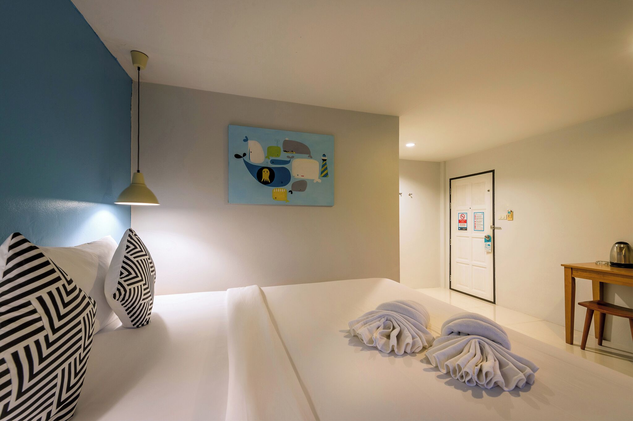 A hotel room with a bed, pillows, a painting, a desk, and a door.