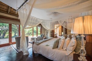 Un dormitorio amplio con una cama grande, una mesita de noche y una lámpara. Hay una ventana con vistas a árboles y un balcón con una silla.