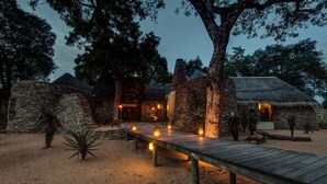 Una cabaña rústica con muros de piedra y techos de paja, una terraza de madera y un sendero arenoso que conduce a la entrada.