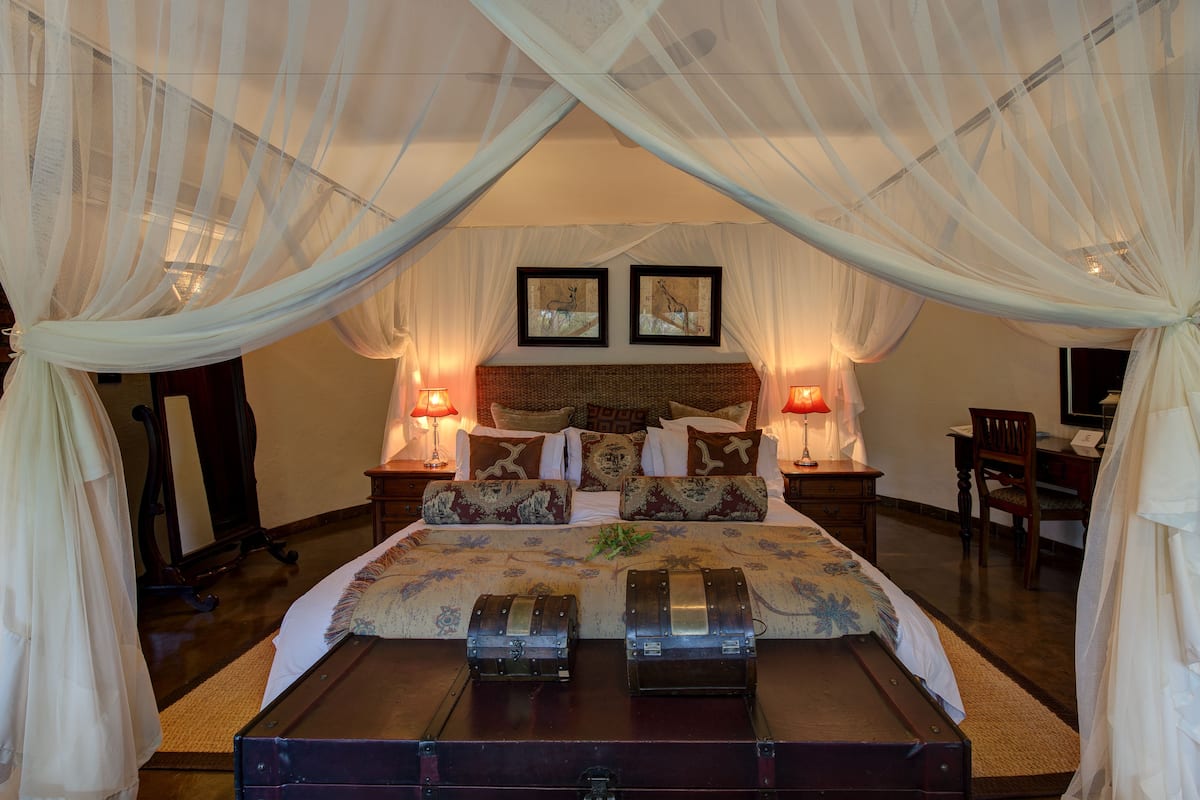 A bedroom with a canopy bed, two bedside lamps, a wooden nightstand, a chair, and two trunks on the bed.