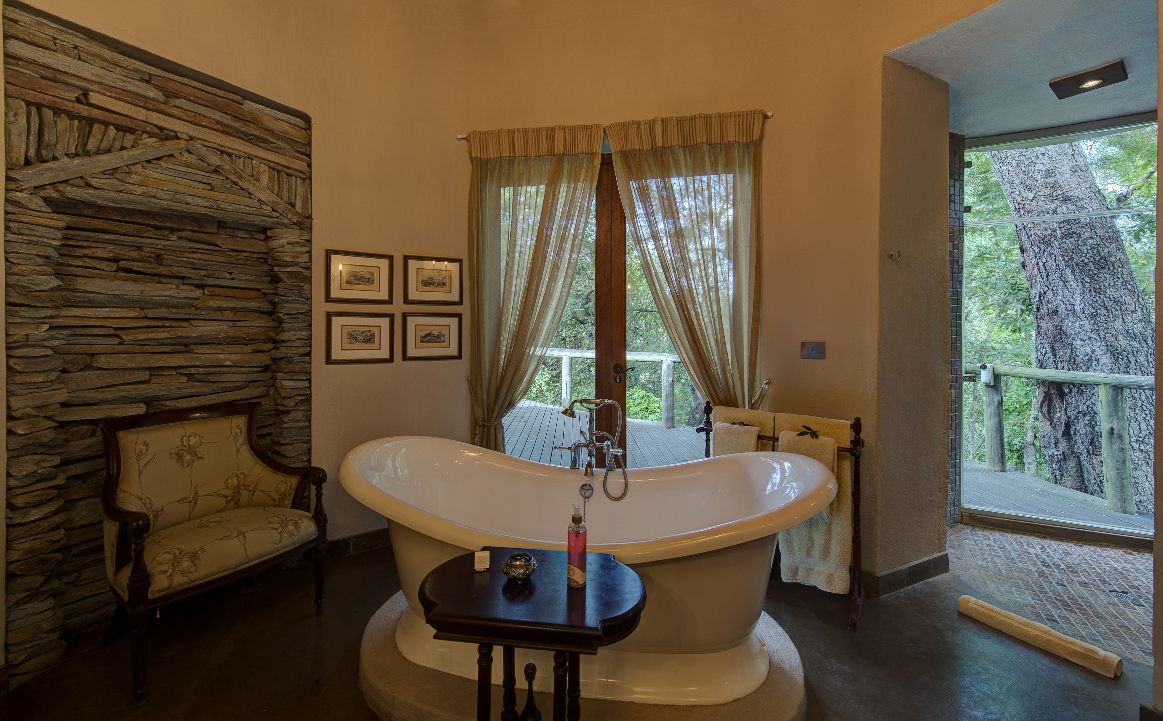 A bathroom with a freestanding bathtub, a wooden bench, stone wall, framed pictures, and a view of trees through large windows.