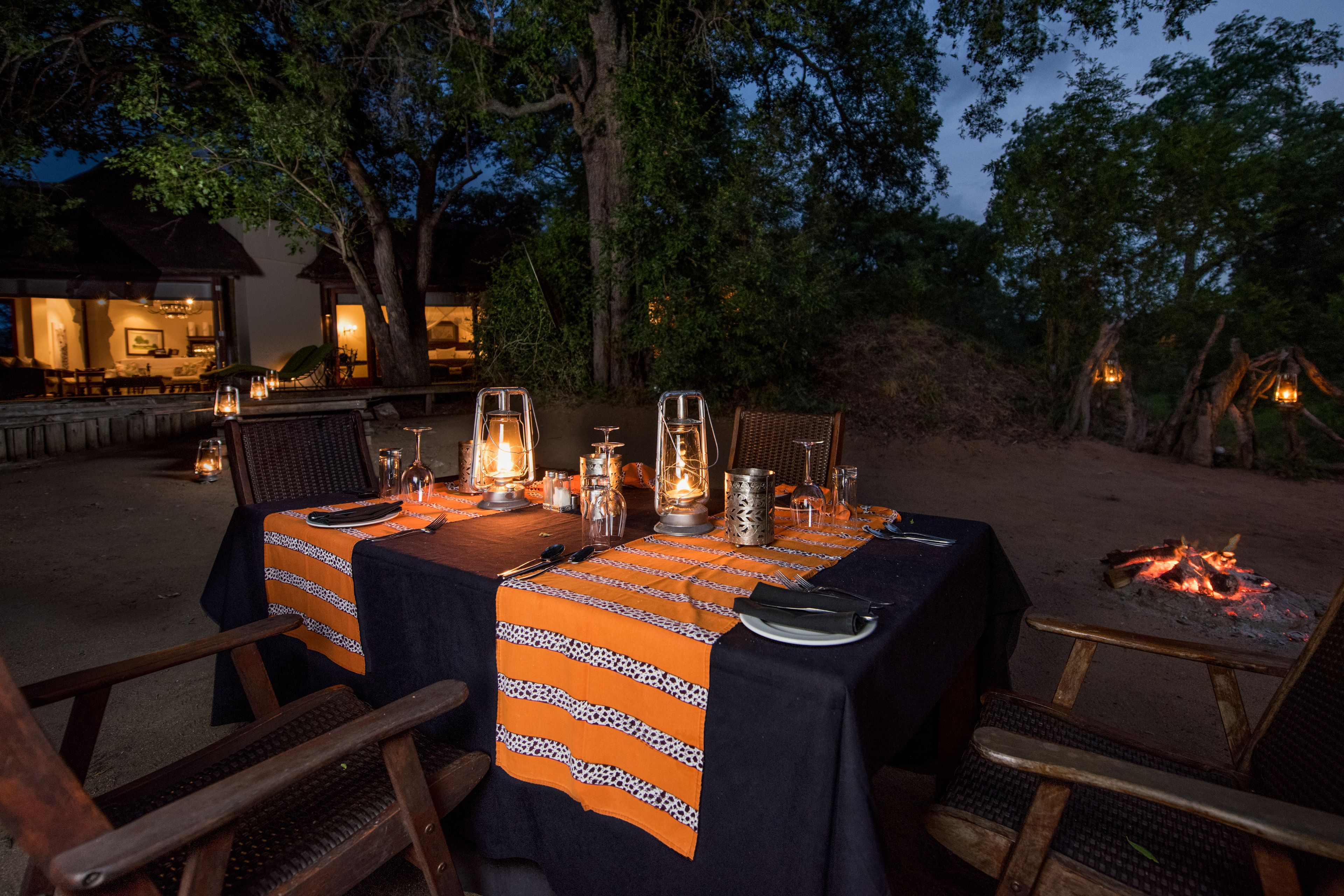 Un área de comedor al aire libre con mesas preparadas para dos personas, rodeada de árboles y un brasero.