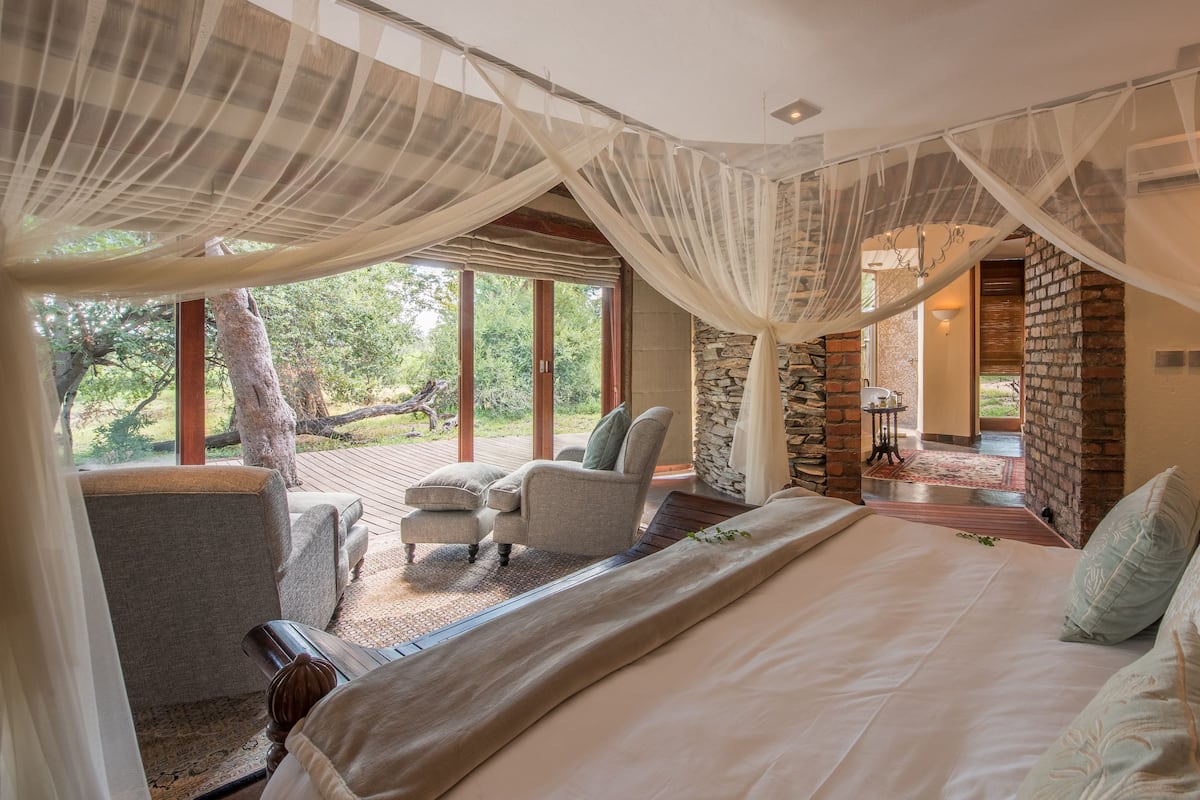 A bedroom with a large bed, a chair, and a small table. There are windows and a view of trees outside.