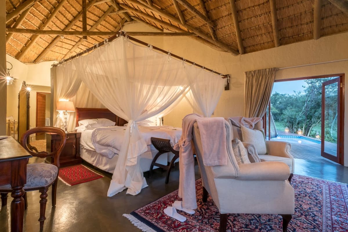 A room with a bed under a canopy, a chair, a table, and a view of the outdoors.