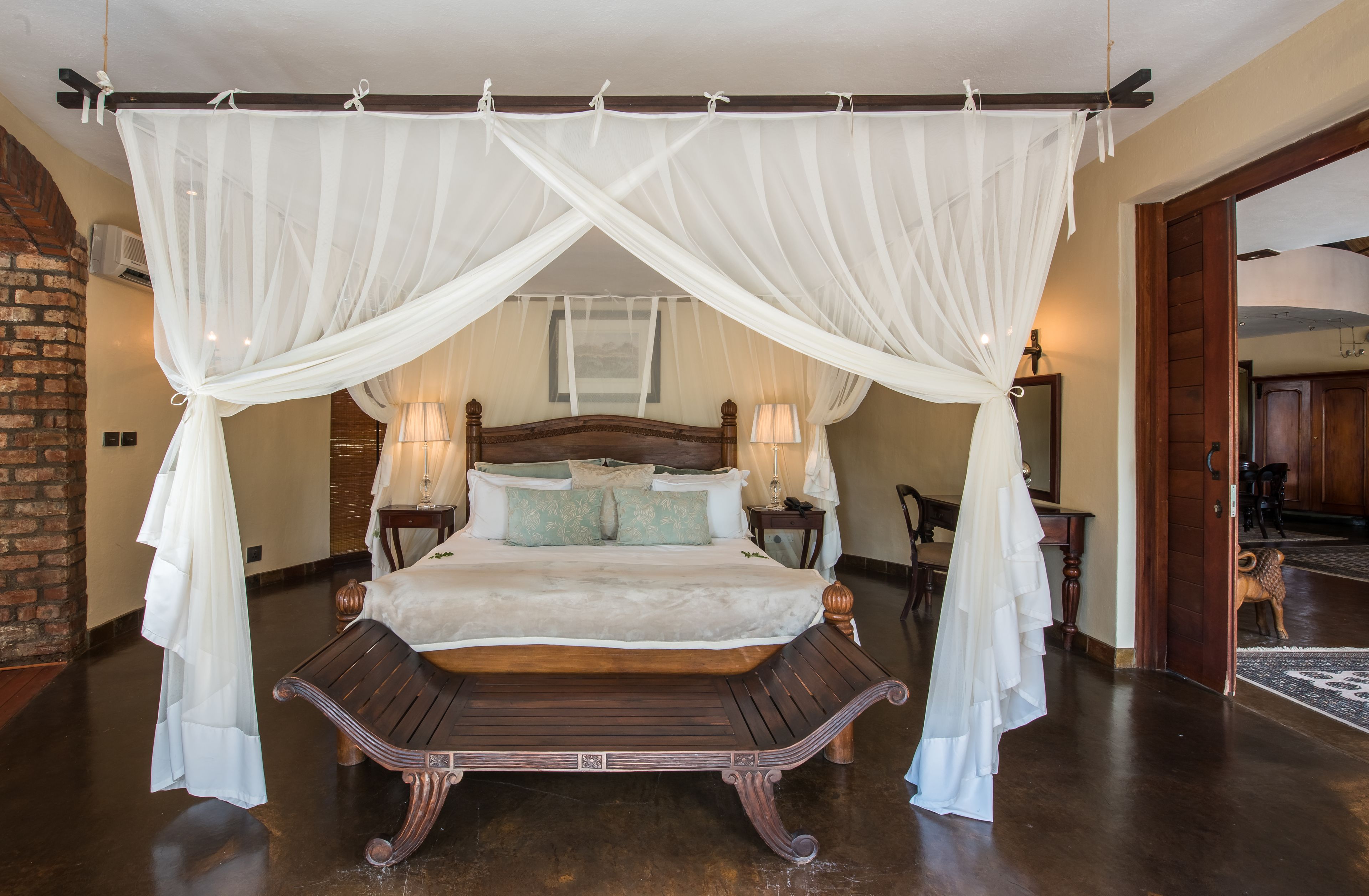 A bedroom with a four-poster canopy bed, wooden bench, two bedside tables with lamps, and a brick wall.